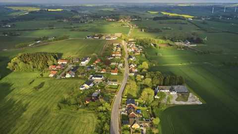 Luftfoto af en lille landsby omgivet af grønne marker og vindmøller i det fjerne.